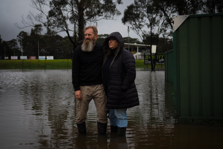 A couple wearing gumboots stand in floodwaters in front of a sports oval