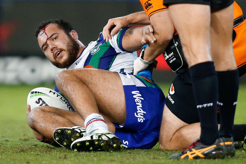 A male NRL player lies on the ground with blood pouring from his forehead as he holds a football.