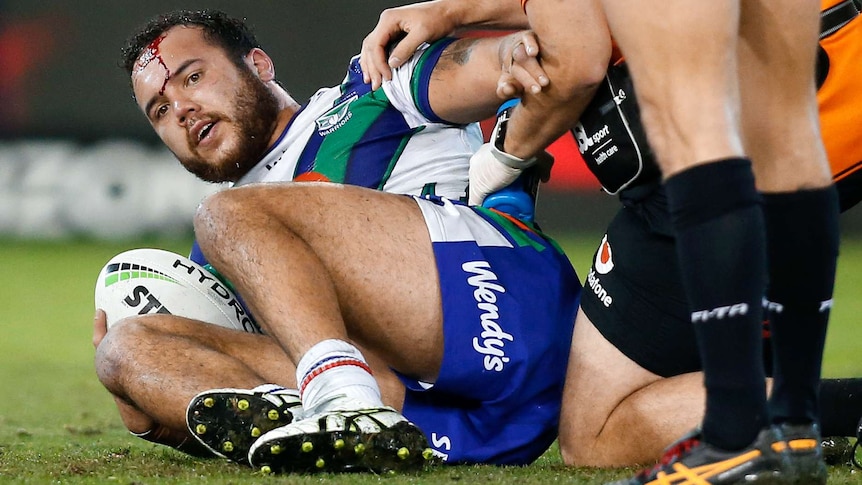 A male NRL player lies on the ground with blood pouring from his forehead as he holds a football.