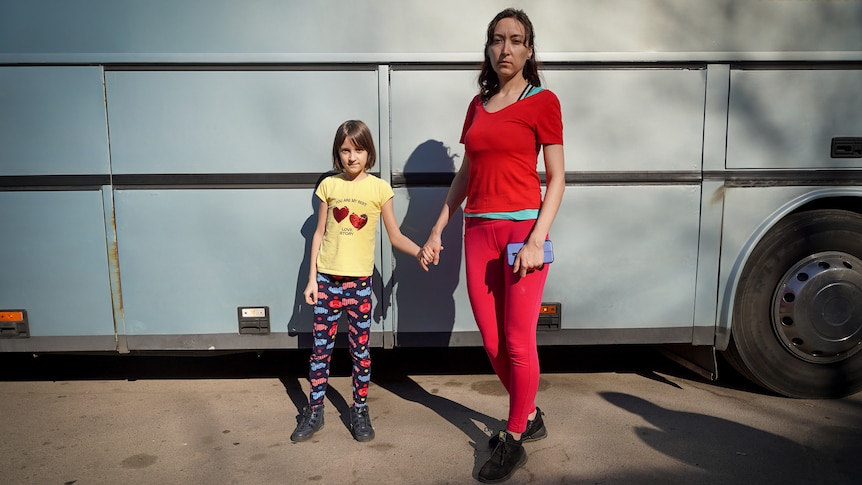 Svitlana stands holding her daugher's hand against the blue backdrop of the bus