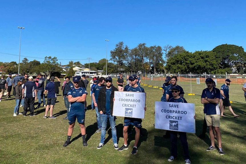 People in blue uniforms holding signs protest outside a fenced-off cricket wicket