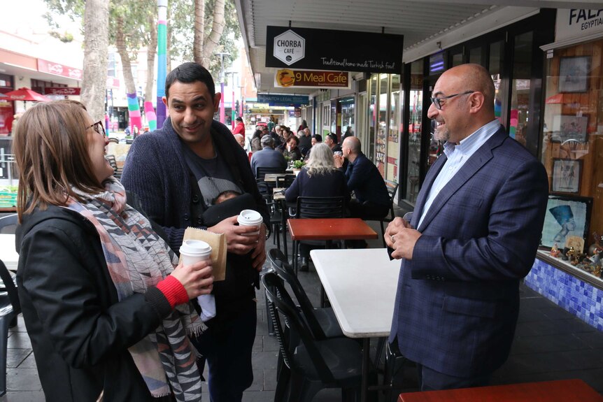 Two men and a woman standing on footpath with coffee cups talking.