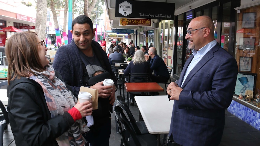 Two men and a woman standing on footpath with coffee cups talking.