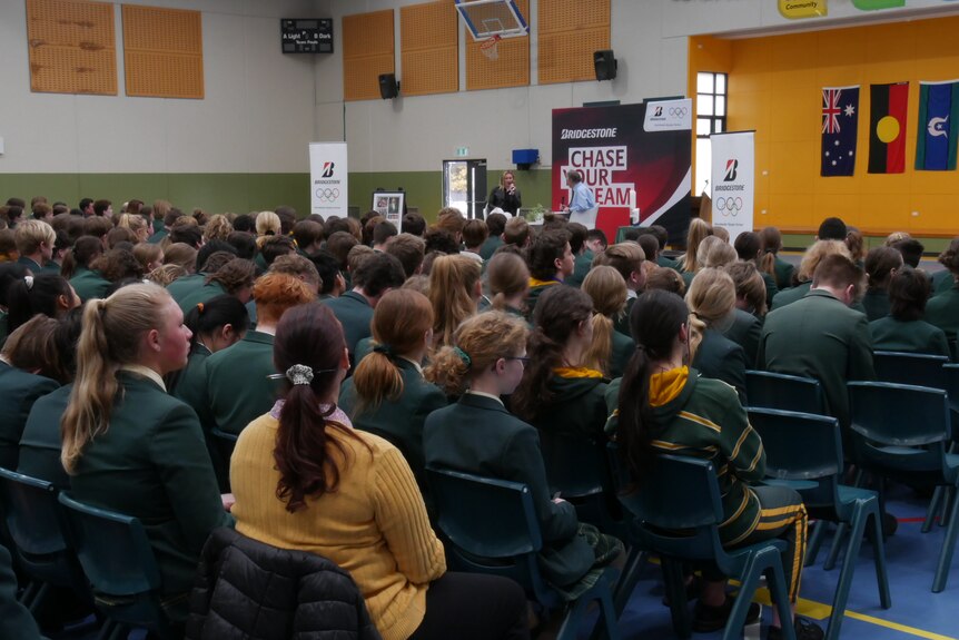 Students in a yellow and green uniform seated in rows to watch Ariarne Titmus speak.