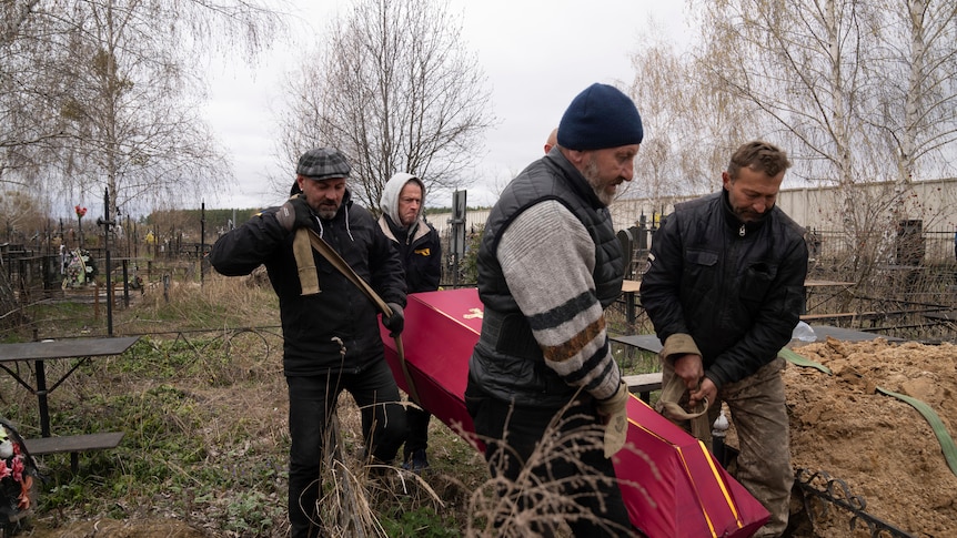 Four men carry a red-coloured coffin through a graveyard on an overcast day