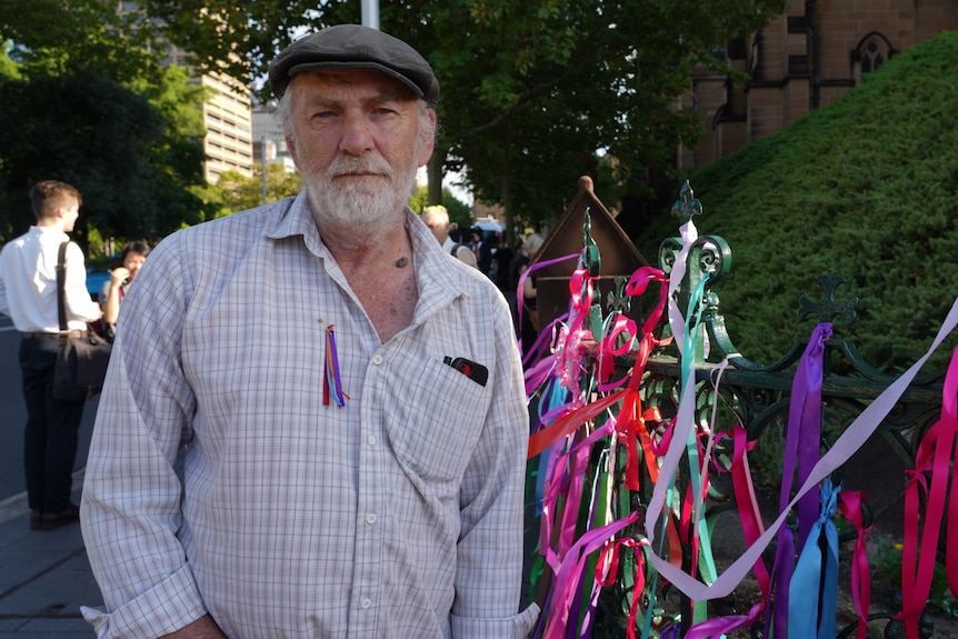 A man wearing a ribbon pinned to his chest stands next to a fence covered in colourful ribbons