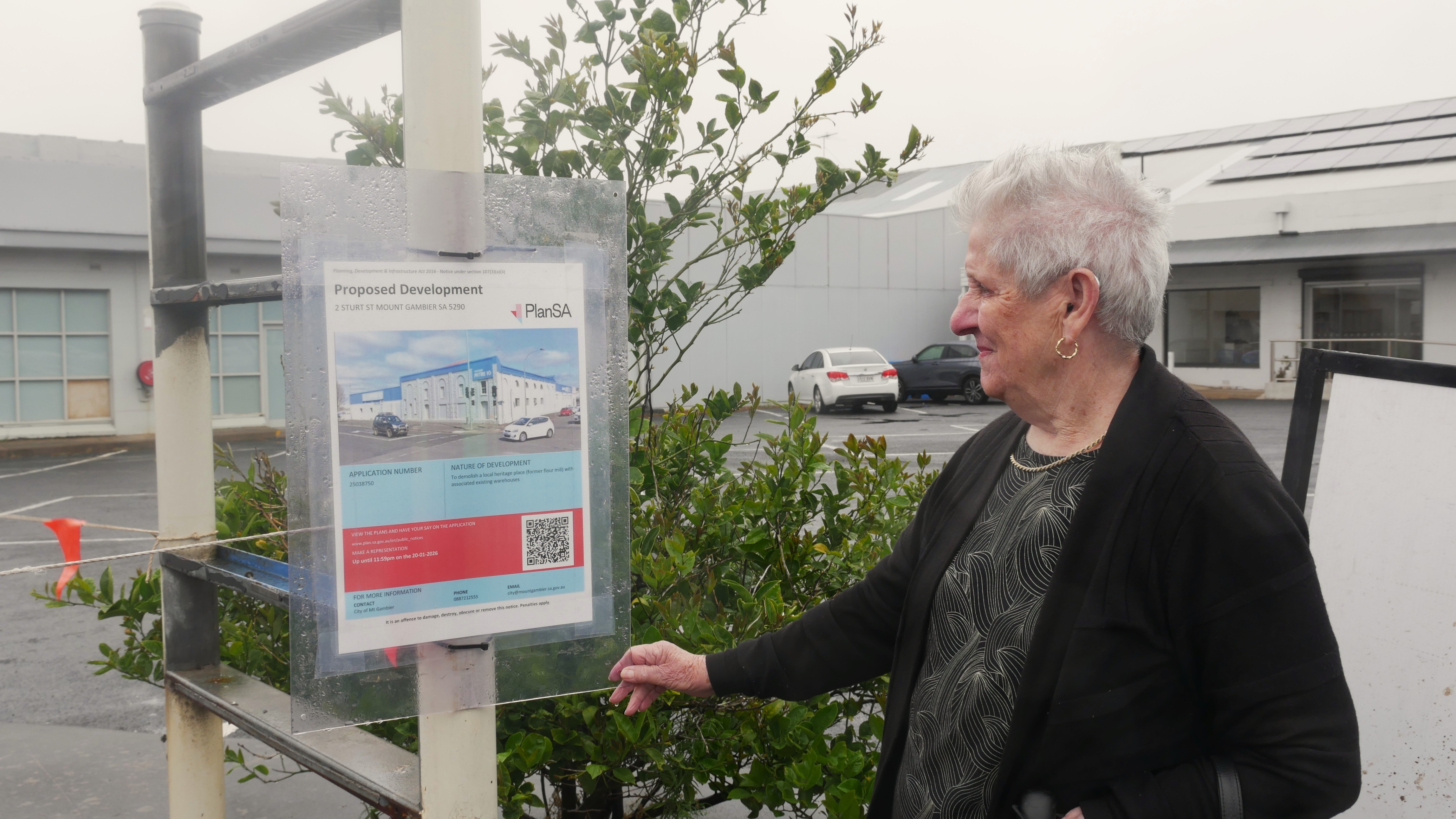 Uma mulher mais velha, de cabelos grisalhos, está olhando para uma placa de papel laminado azul e vermelho do lado de fora de um estacionamento em um dia chuvoso.