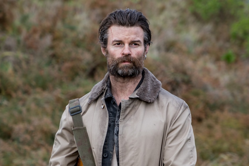 A 40-something man with cropped hair and a thick beard stands in the New Zealand countryside with a dazed expression