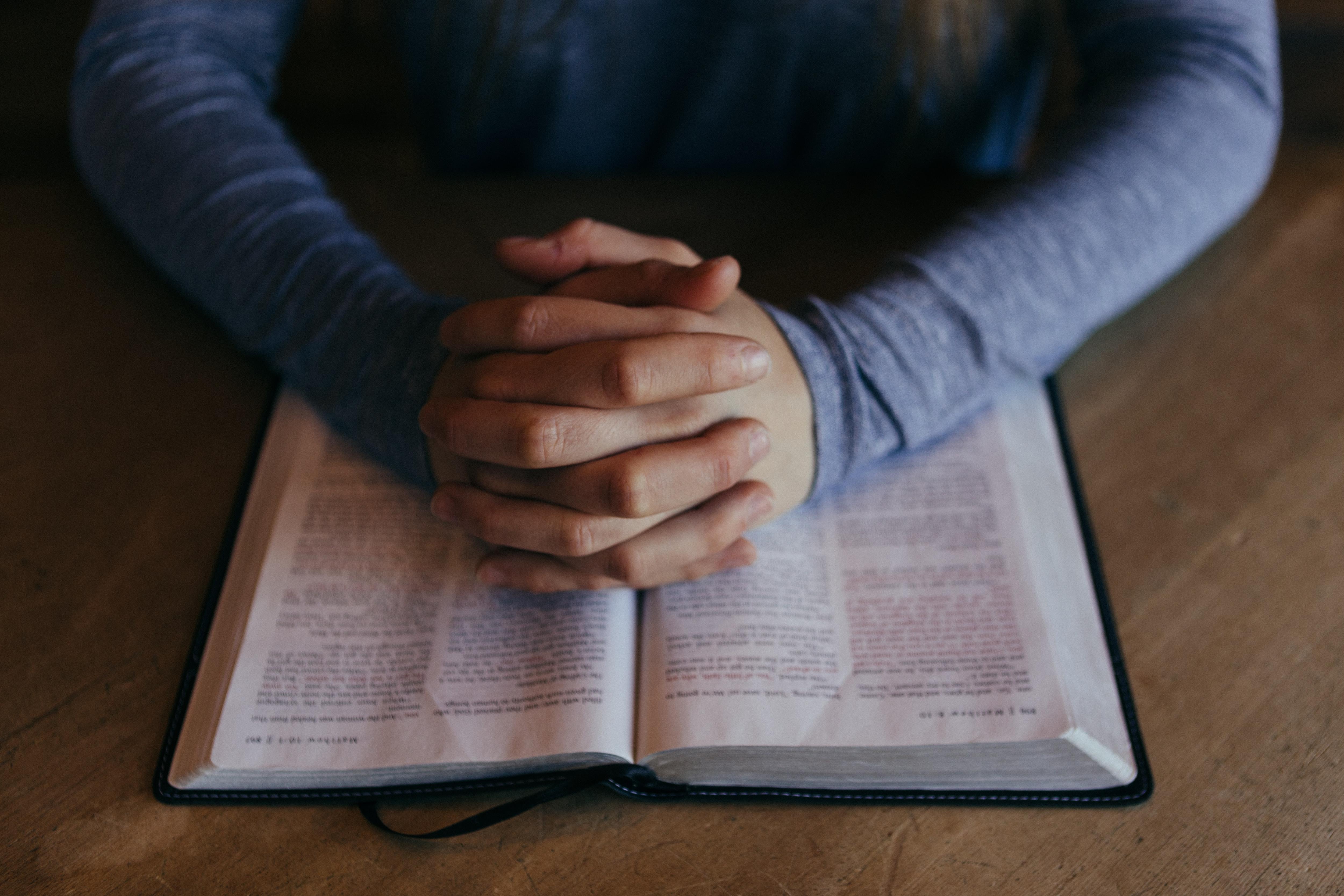 Close-up of hands clasped as if in prayer over an old book with blurred text, which could be a religious text.