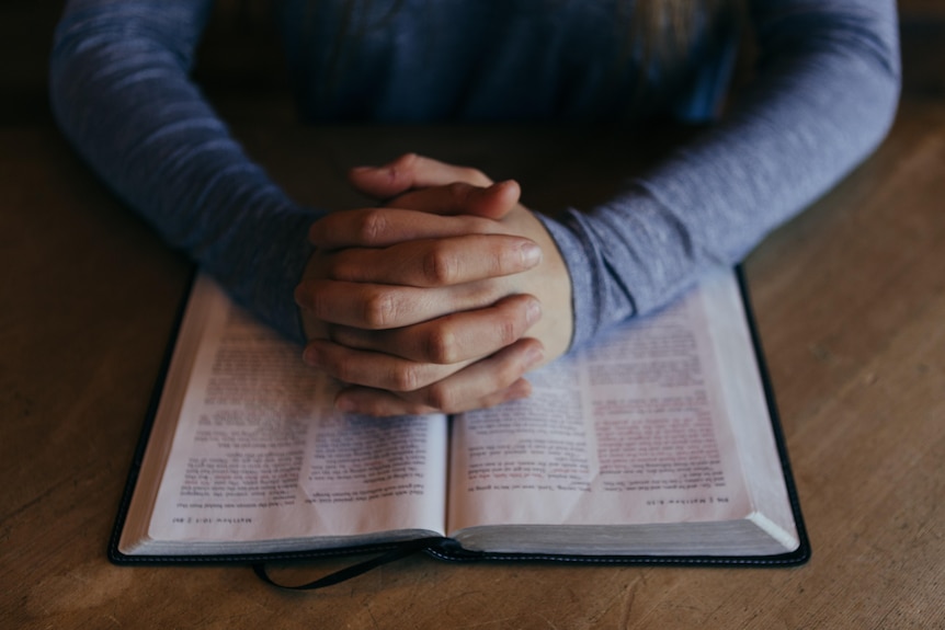 Close-up of hands clasped as if in prayer over an old book with blurred text, which could be a religious text.