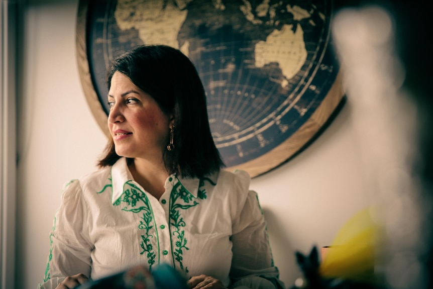 A woman with shoulder-length hair stands in front of an atlas, looking over her right shoulder