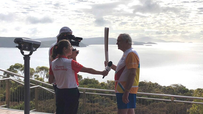 A man, interviewed by two people, carries the Queen's Baton with the sea and sky of south-west WA in the background