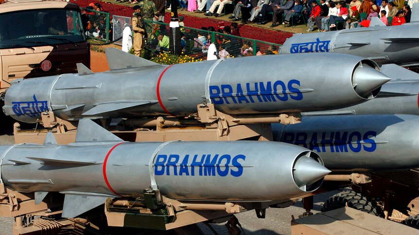 Several missiles with blue writing "BrahMos" are being paraded down a road. 
