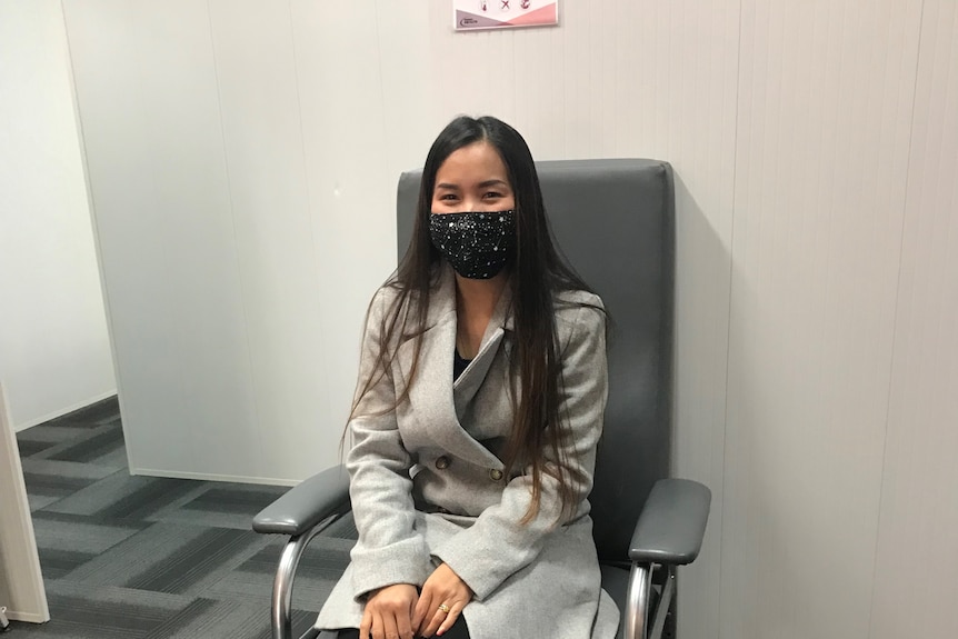 A woman in a grey jacket, wearing a face mask, sits on a chair waiting for her vaccination. 