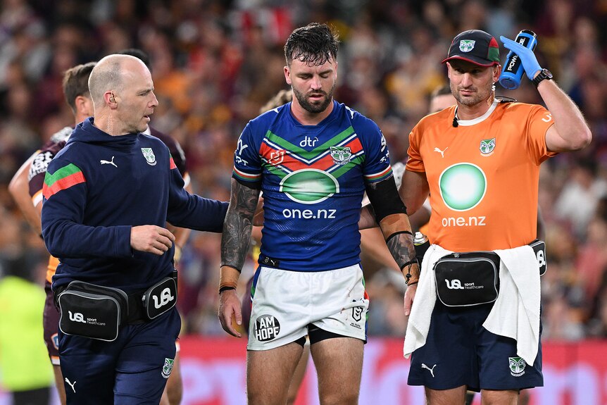 An NRL player, looking groggy, is escorted from the field by two male trainers, one signalling to his head.