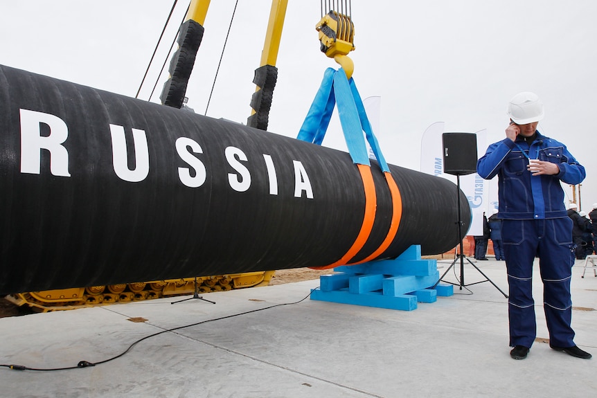 A man on the phone stands in front of an industrial pipe labeled "Russia"