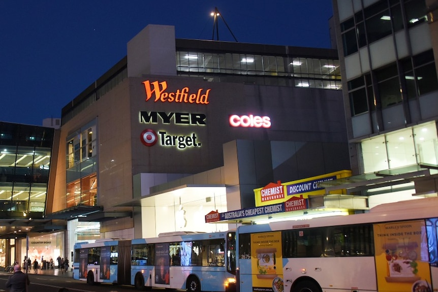 A Westfield shopping centre advertising several shops at night with buses in front 