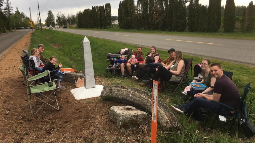 A photo shows a group of people gathered at the roadside on camping chairs, separated by a ditch