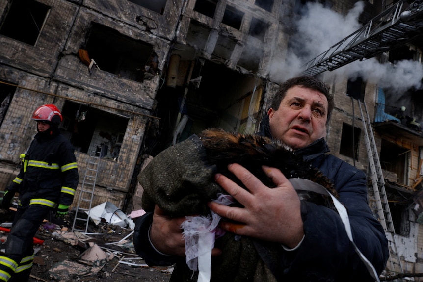 A man carries his belongings in his arms with his burntout home seen behind him.