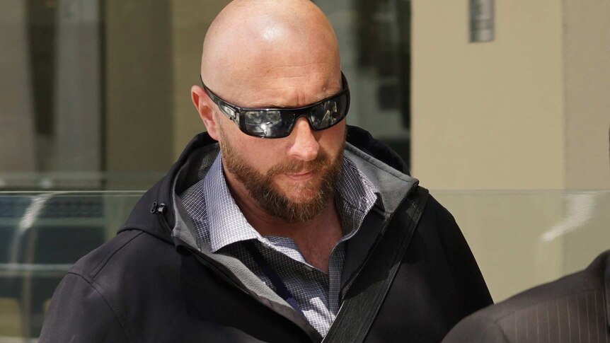 A bald man wearing a black jacket and sunglasses walks along the street.