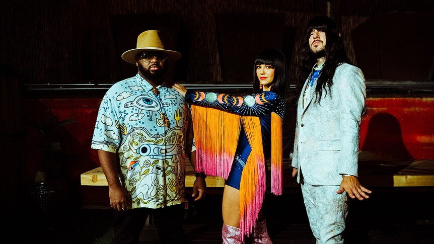 Three members of the band Khruangbin stand side-by-side wearing brightly coloured clothes and looking into the camera.