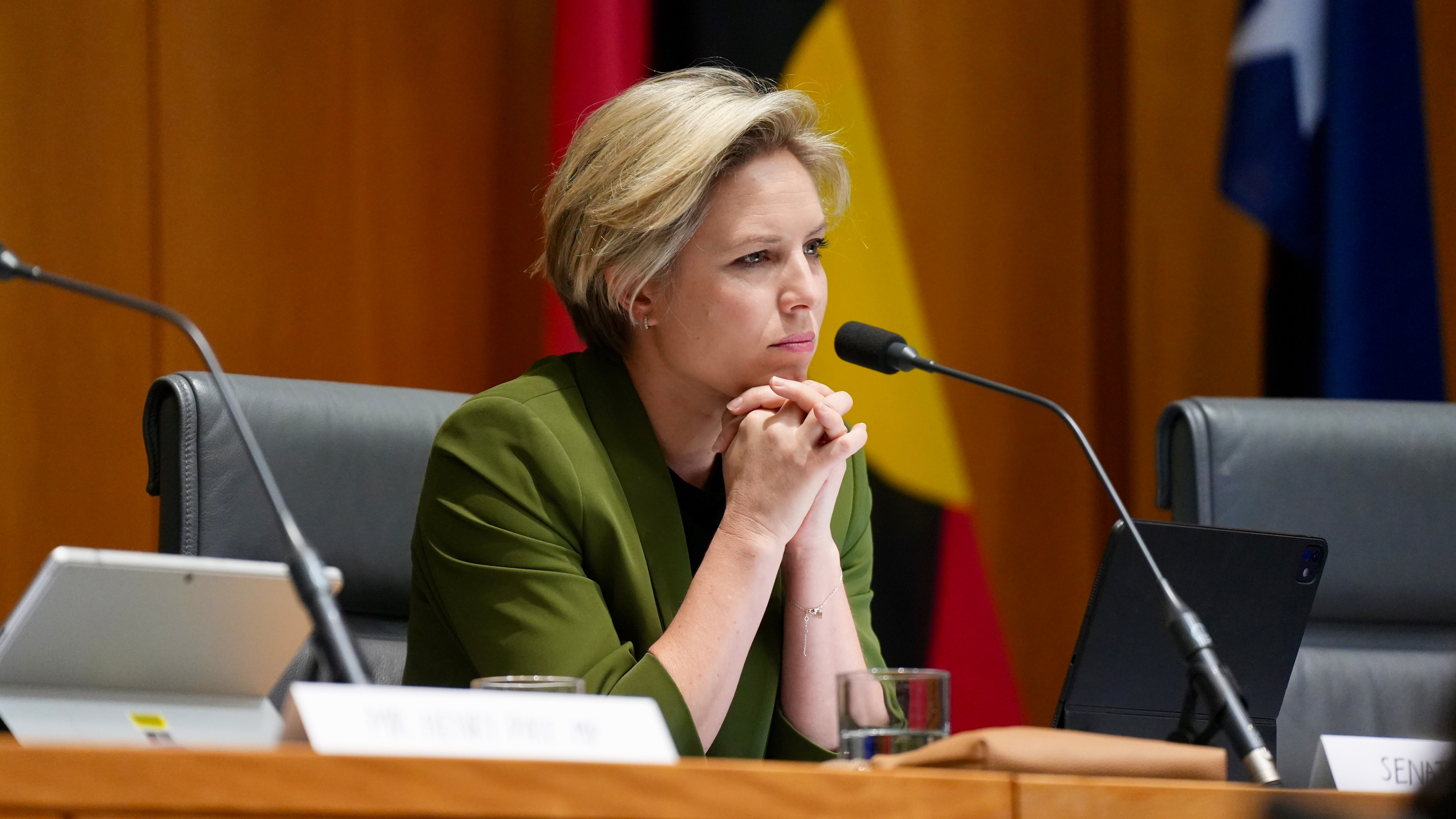 A woman with a blonde bob, in a green shirt, sits at a Senate inquiry desk.