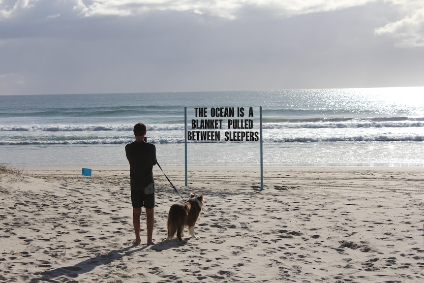 Man with dog on beach looking at sculpture with writing 'The ocean is a blanket between sleepers'. 