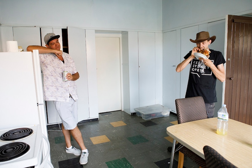 A man leans on a kitchen fridge while another bites into a whole roast chicken for a story on share housing.