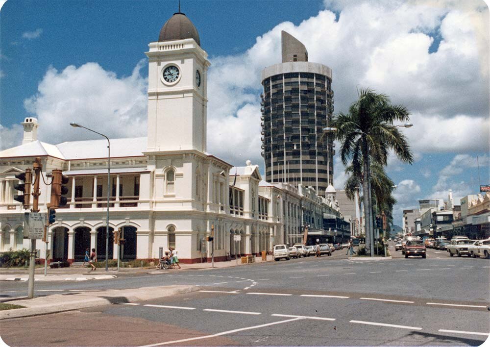 An old, but color photograph of a busy city street.  A post office sits before a much taller circular high rise building. 