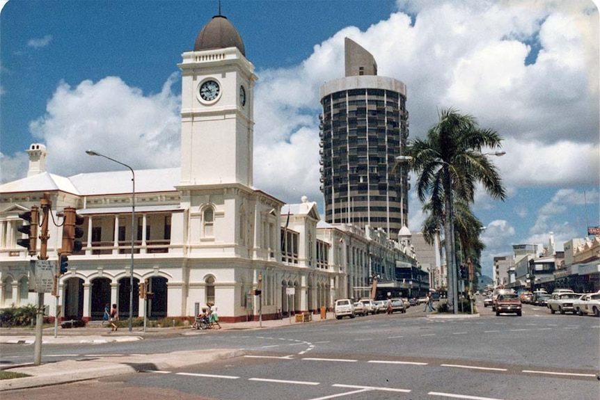 An old, but color photograph of a busy city street.  A post office sits before a much taller circular high rise building. 