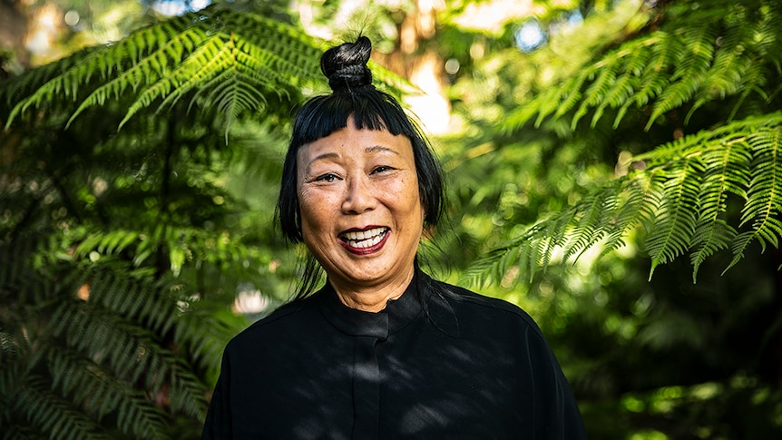 A woman with black hair tied up in top knot and Mao collar style jacket laughs in lush green outdoor area surrounded by ferns.