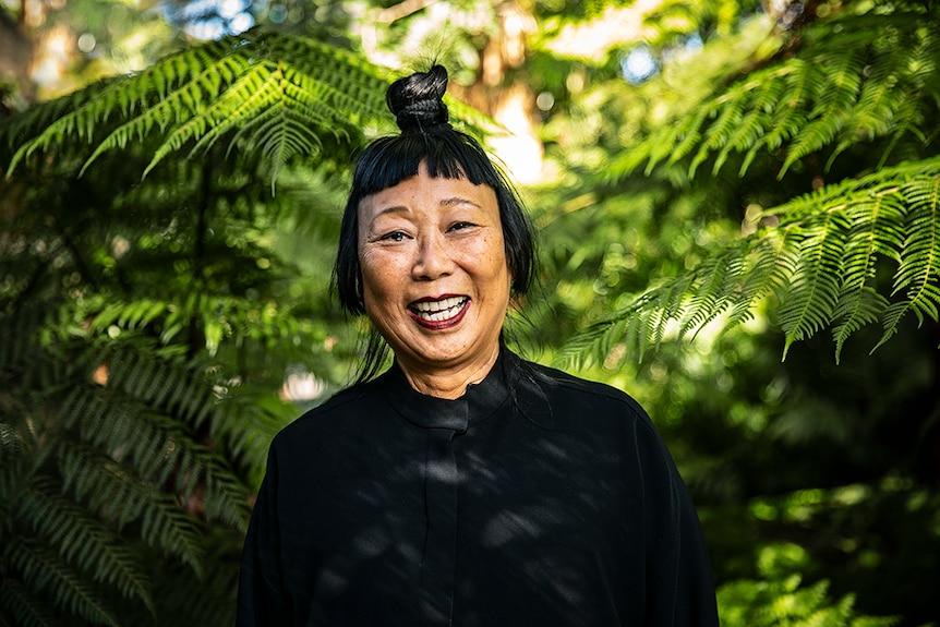 A woman with black hair tied up in top knot and Mao collar style jacket laughs in lush green outdoor area surrounded by ferns.