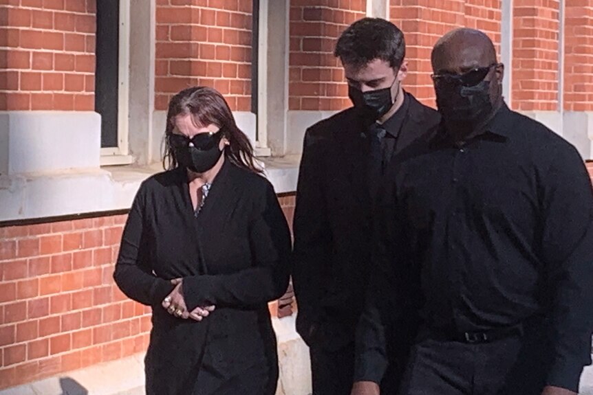 Un jeune homme marche la tête baissée portant un masque facial flanqué d'une femme et d'un homme chauve, tous deux portant des lunettes de soleil.