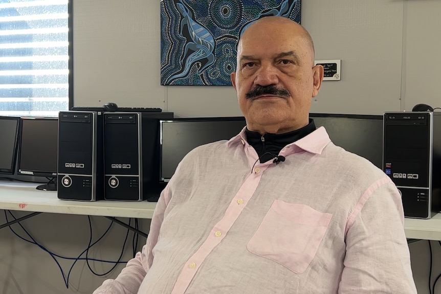 A man in a button-up shirt with a moustache sitting in a chair with computer monitors and Aboriginal artwork behind him.