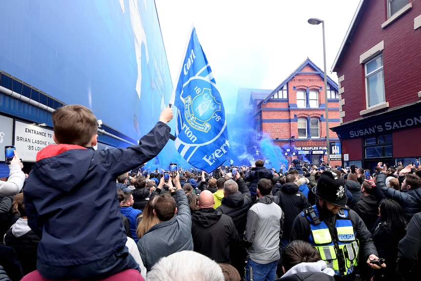 A child flies an Everton flag as blue smoke sails above a crowd of fans outside Goodison Park