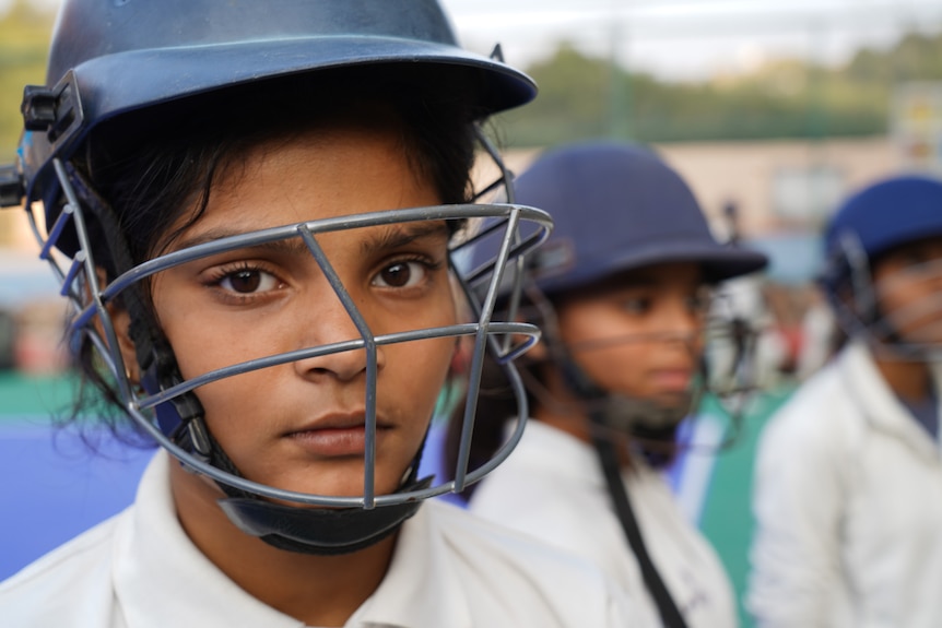 A close up of a girl with brown eyes wearing a cricket helmet and uniform while standing with similarly dressed girls.