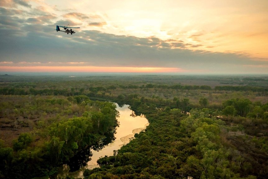 The silhouette of a small aircraft flies over lush green trees and a winding river below a pink, blue and yellow sunset.