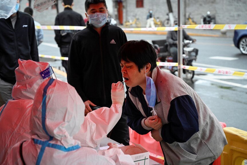 Азиатская женщина с короткими волосами наклоняется к медицинскому работнику в защитном костюме, чтобы взять мазок изо рта.
