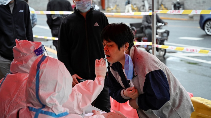 Asian woman with short hair bends towards health worker in hazmat suit to receive mouth swab