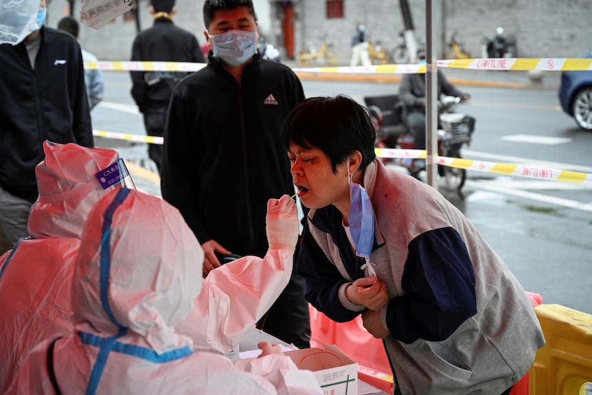 一名短发的亚洲女性弯身向一名身穿防护服的卫生工作者接受口腔拭子