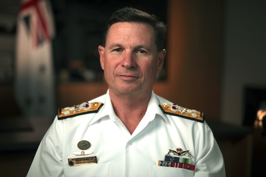 A man sits indoors in a white navy uniform looking at the camera. Behind him hangs a white Australian Navy flag.