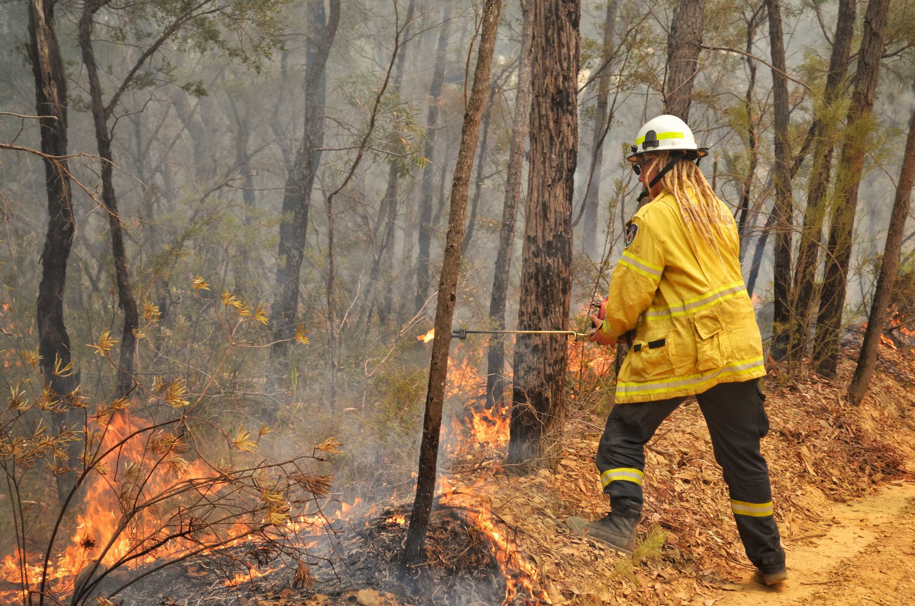 The firefighter has blonde dreadlocks, and is holding a firelighter as scrub and trees burn in front of her.