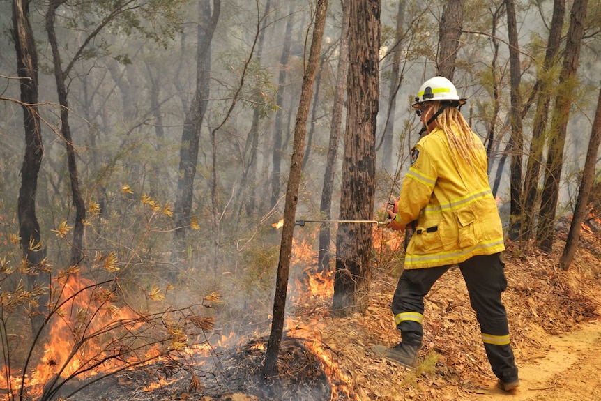 The firefighter has blonde dreadlocks, and is holding a firelighter as scrub and trees burn in front of her.