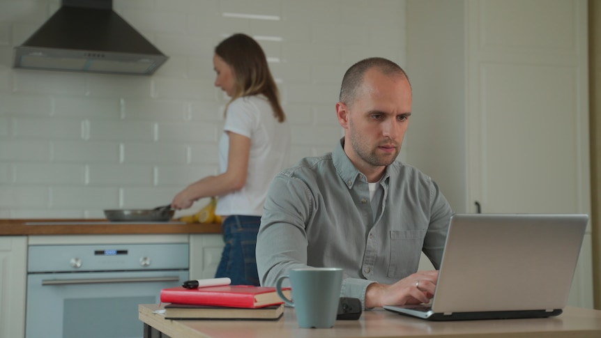 Man in grey using laptop on kitchen table with woman at stove in background. Coffee cup on table