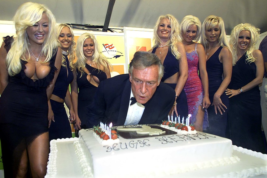Hugh Hefner leans over a giant birthday cake as he blows out candles, surrounded by seven blonde women.