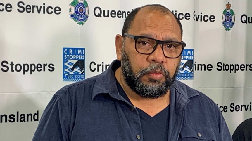 A man in a blue shirt and glasses stands behind two microphones in front of a queensland police banner