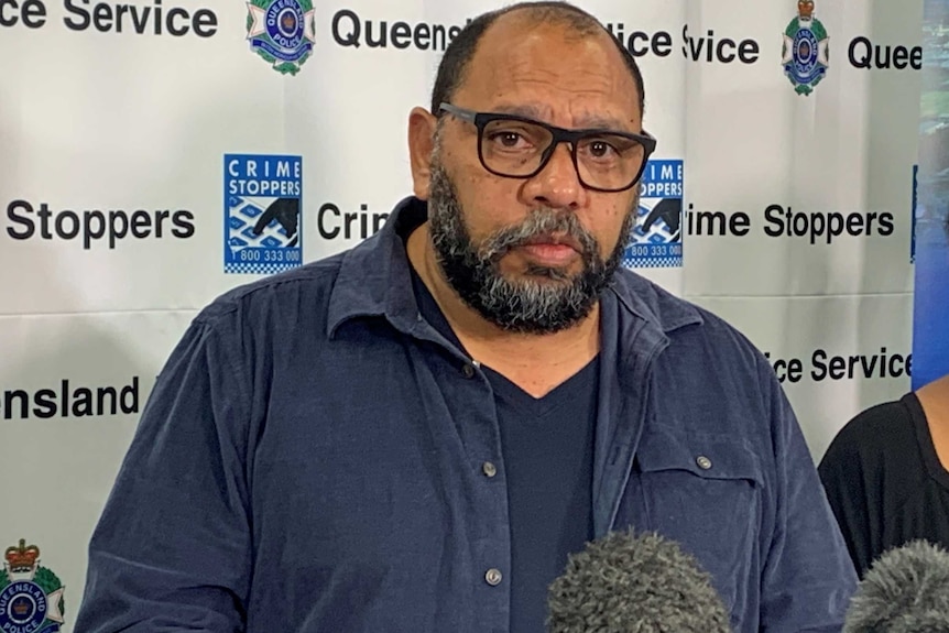 A man in a blue shirt and glasses stands behind two microphones in front of a queensland police banner