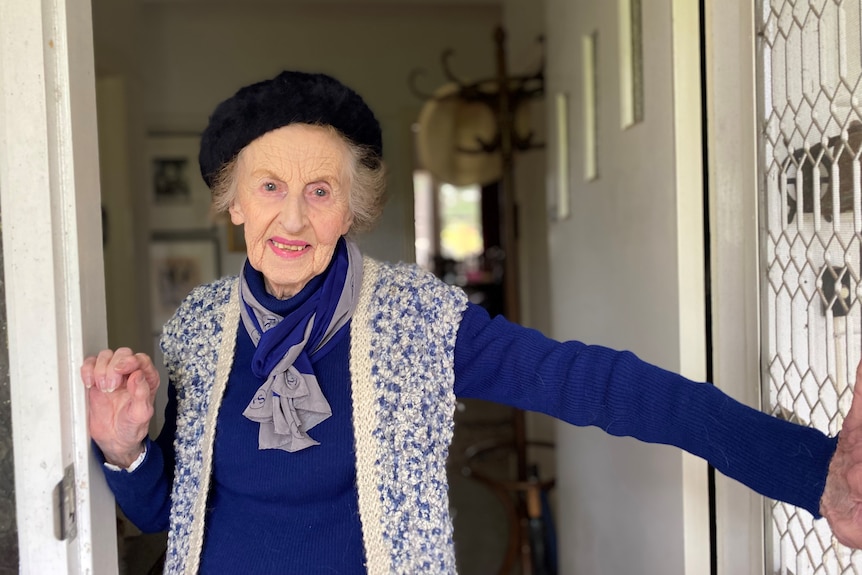 An elegant elderly woman dressed in blue woollen dress matching scarf and knitted shawl with beret stands in doorway