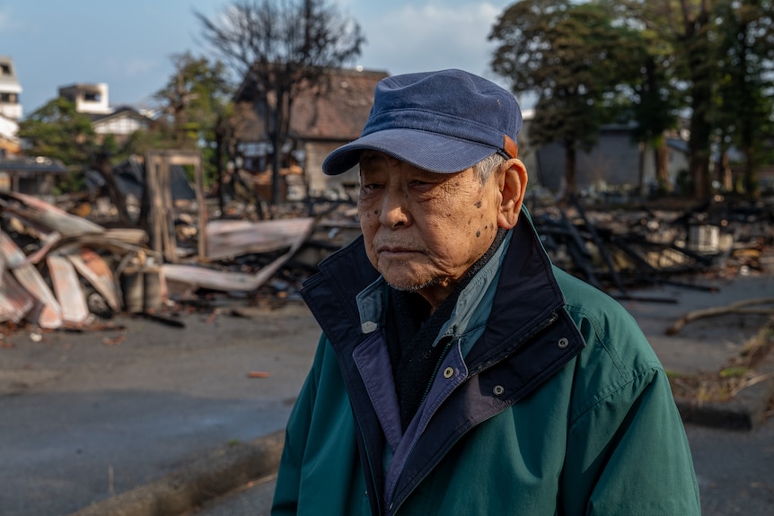 Image of an old man wearing a blue hat and a green raincoat. Behind him are houses that have been destroyed.