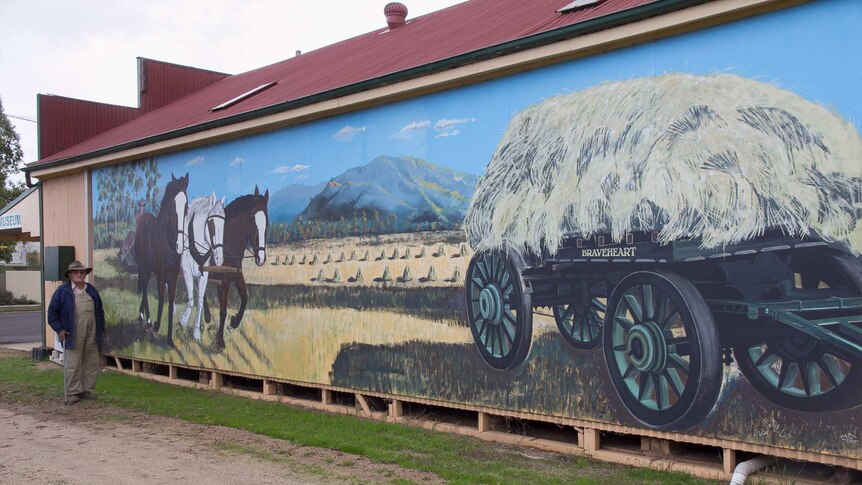 An old man in hat and walking stick in front of a mural on a building showing a paddock being ploughed with horses
